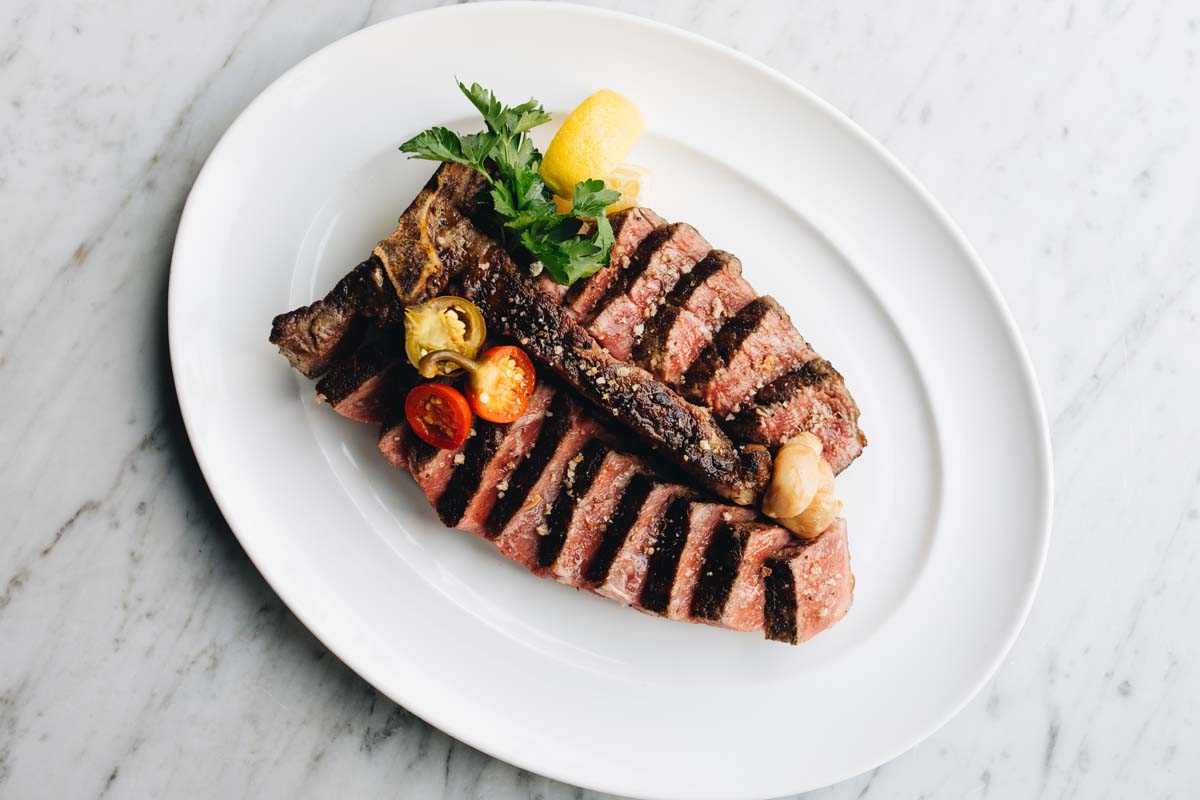 Overhead shot of a t-bone steak pre-cut with cherry peppers, lemon wedges, and parsley to garnish