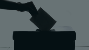 A backlit photo of a hand putting a ballot in a box.