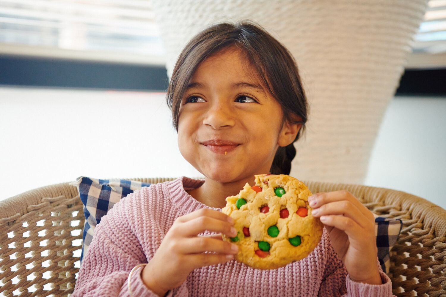 A girl smiling while holding a cookie sitting on a wicker chair.